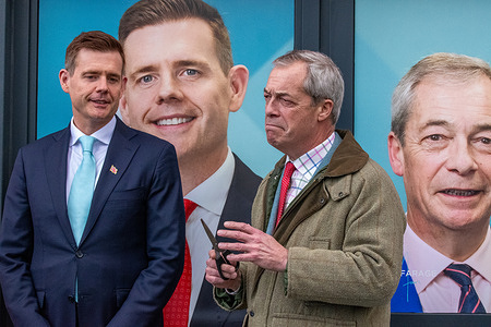 Nigel Farage seen holding scissors to cut the ribbon and Reform UK candidate Matt Goodwin, during the campaign. Nigel Farage’s campaign visit to Gorton and Denton where he opened, by cutting a light blue ribbon, the Campaign HQ with Reform UK candidate Matt Goodwin. They were joined by Reform MP, for Runcorn and Helsby, Sarah Pochin who had to step over the ribbon before the cutting. Reform UK supporters held signs and waited in the cold. After the official opening Nigel Farrage and Matt Goodwin answered questions from the press together.