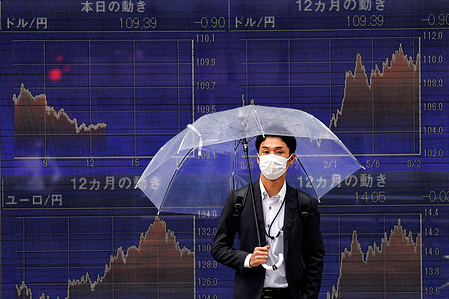 A man holding an umbrella walks in front of an electric board showing Nikkei index a brokerage in Tokyo.