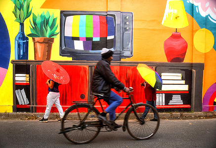 A cyclist with his bicycle seen passing in front of the vibrant Colour street's of Kolkata named as Kolkata's "Art Corridor" . A street is decorated with various wall art depicting the daily household life of a Bengali household in Kolkata, India . The project named as Kolkata's "Art Corridor" primarily refers to the vibrant Colour Corridor street art project which is transforming a laneway into a massive mural using over 5,300 shades to explore "home away from home" through colour, memory, and Kolkata's culture with artist Sayan Mukherjee in collaboration with Start India Foundation and Asian Paints.