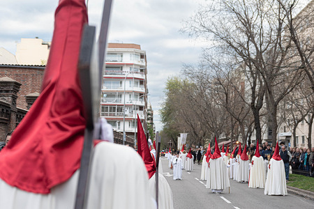Nazarenes dressed in white robes and red pointed hoods walk in formation during a Holy Week procession. The Holy Week procession in Madrid, is a deeply rooted Catholic tradition held during the week leading up to Easter. Featuring solemn parades with religious floats, traditional music, and participants in penitential robes, the event reflects Spain’s rich spiritual heritage and draws thousands of spectators each year.