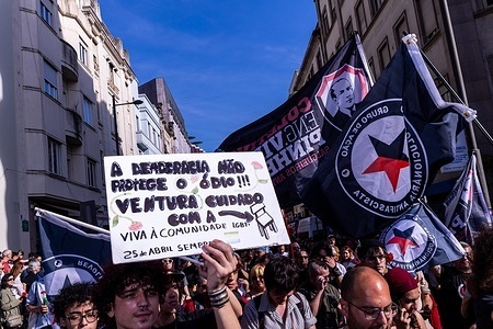 A crowd marches holding antifascist flags and a placard with political messages during the rally. Thousands of people marched through several Portuguese cities during the commemoration. Demonstrators carried red carnations, banners and political messages referencing democracy, social rights, antifascism, education, housing and public services. The marches included families, unions, activists, students and community groups. The events took place in urban streets and public squares, with participants highlighting the ongoing relevance of the revolution’s values.