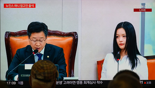 A 24-hour Yonhapnews TV broadcast at Yongsan Railway Station in Seoul showing a live news broadcast with Hanni (R), a member of the girl group NewJeans, testifies as a witness about workplace bullying directed at her during an inspection by the labor and environment committee at the South Korea's National Assembly in Seoul. NewJeans' Hanni attended a parliamentary labor committee meeting addressing allegations of her workplace, bullying within Hybe, the parent company of ADOR, NewJeans' label.