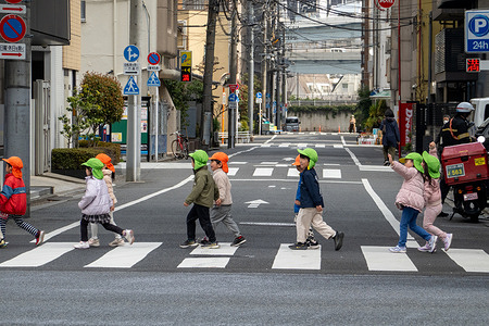 Young children wearing colorful safety hats cross a street with a caregiver in central Tokyo, Japan. The hats are commonly used by preschools and daycare groups to keep children visible during outdoor walks. Central Tokyo is a dense urban core where business districts, shopping areas, and entertainment neighborhoods intersect. It combines modern skyscrapers, historic sites, and major transit hubs, reflecting the city’s fast-paced and layered character.