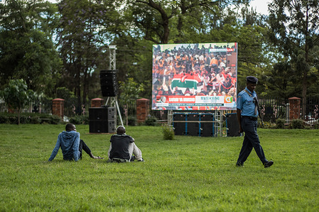 A police officer walks past a group of people following the memorial service proceedings of the former President of Kenya on a large screen at Nyayo Garden. The late President Mwai Kibaki was the third president of Kenya after the Late Jomo Kenyatta and Late Daniel Moi. He rose to power in December 2002 until April 2013 when his second term in office came to an end. He has been named as the most progressive president Kenya had.