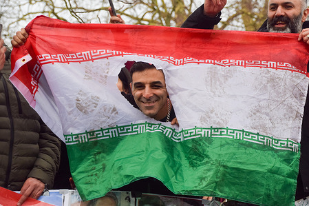 A protester puts his face through a hole in a defaced flag of the Islamic Republic of Iran as pro-monarchy Iranians celebrate the death of Khamenei outside the Embassy of Iran and call for Reza Pahlavi to take over as King of Iran. Ayatollah Khamenei was killed in air strikes by USA and Israel.