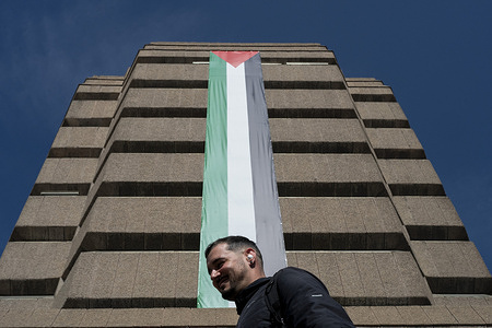 A man walks beneath a large Palestinian flag hanging from a building, this low-angle shot captures a public display of solidarity and political expression in an urban setting.