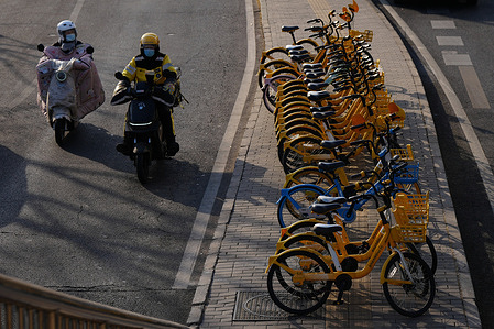 Two people on electric scooters ride past a large number of shared bikes parked on the roadside.