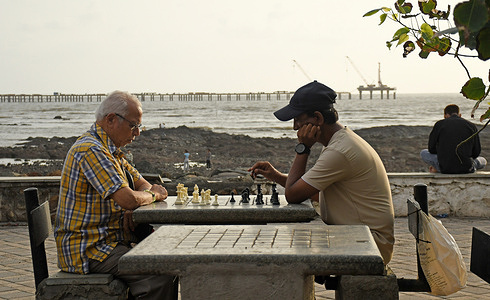 People are seen playing chess at a sea facing promenade. Young and old alike enjoy playing a game of Chess at the open air picturesque sea facing promenade where Chess boards are set in marble stone tables.