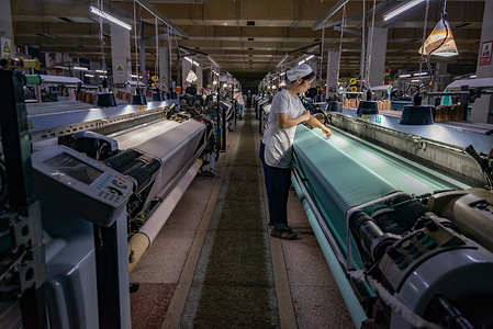 A worker works on a machine with turquoise silk in a factory aisle. Anhui Jingjiu Silk Co. Ltd. is a comprehensive cocoon and silk enterprise whose main products include raw white silk, real silk satin, and home textile clothing.