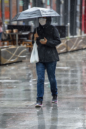 A women wearing a face mask as a precaution against the spread of covid-19 uses an umbrella during a heavy downpour of rain while other people take shelter near a shop door in London’s Leicester Square.