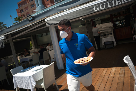 A waiter wearing a face mask is seen serving a plate of prawns to costumers at the terrace of 'Gutierrez' beach bar during the partial lockdown in all the country. Spain is going through a plan of down-scaling towards a "new normality" by relaxing measures that resulted from the COVID-19 outbreak. During phase 1 is allowed that people can visit their families and meet with number maximum of 10 people at homes, outdoors places and terraces. Also can reopen church, business, stores and bars or restaurants with their terraces limited at 50% continuing keeping safety distance and the use of face masks.