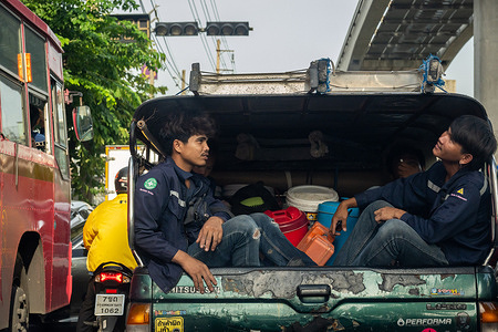 A close-up of two young workers sitting on the back of a truck, with one leaning his head out to watch the city scenery during a traffic jam on Lat Phrao Road in Bangkok. Bangkok's traffic is notoriously congested, especially during rush hours, due to high vehicle density and limited road space. The city relies on numerous bridges to connect districts and alleviate traffic flow across rivers.