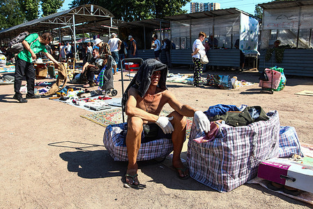 A bare-chested male seller sits with bags on a hot summer day, covering his head from the sun with a jacket at the Udelnaya flea market in St. Petersburg.
