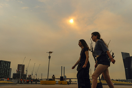 Women seen walking as the red sun casts an orange glow over Docklands with smoke from bushfires visible in the sky. Smoke from multiple bushfires and grassfires burning the city is affecting air quality and visibility, turning the sun red including the CBD. The Department of Environment, Land, Water and Planning (DELWP) has issued a community information message advising that smoke and ash may be seen or smelled in areas far from the fires. Residents are urged to take precautions especially those with respiratory conditions as bushfire activity continues.