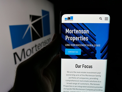In this photo illustration, a person is holding a smartphone with the web page of construction business M.A. Mortenson Company on screen in front of logo.
