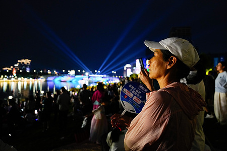 People stand on the lawn enjoying a water concert at the Canal Park. Canal Park in Beijing hosted an outdoor water concert, featuring live musical performances set against the park's scenic Grand Canal backdrop. Attendees gathered along the surrounding riverside lawns and waterfront walkways to enjoy the open-air summer music event.