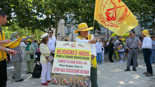 A demonstrator holds a large yellow flag of the People's Mojahedin Organization of Iran (MEK) next to a large poster reading "Rejoignez-nous pour la grande manifestation... Stop aux exécutions en Iran" (Join us for the grand demonstration... Stop executions in Iran). Demonstrators gathered at Place de la République and near Place Vauban to participate in the "Free Iran 2026 Onward to a Democratic Republic" rally. Defying a late ban imposed by French authorities, protesters carried Iranian flags featuring the Lion and Sun and held placards demanding democracy.