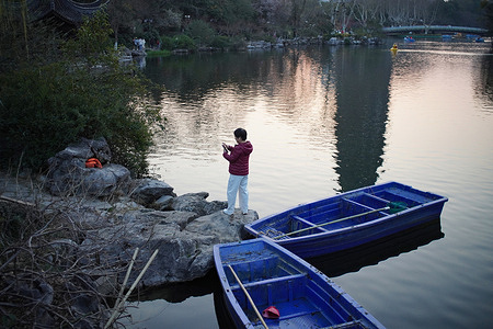 A woman takes photos of the tranquil lake and blue rowboats at dusk.