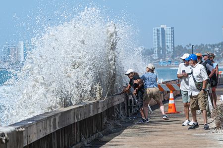 A woman seen trying to protect an old man by pulling him away as the crashing wave hits the seawall. Large swells driven by Hurricane Polo slammed into the Southern California coastline, causing elevated tides and high surf in Long Beach. Powerful waves crashed against seawalls and boardwalks on the Long Beach Peninsula, threatening nearby beachfront homes and prompting city crews to fortify the shoreline with emergency sandbags.
