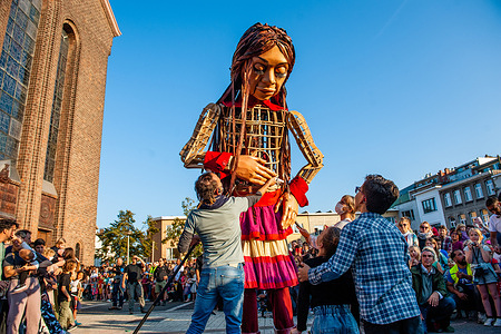 Two siblings are seen giving a letter to Little Amal. 
A giant puppet is currently grabbing attention across Europe to the plight of young refugees who have fled from Syria. Little Amal represents a nine-year-old Syrian refugee girl and is over 11-feet-tall. She has been walking nearly 8,000 km from the Syrian-Turkish border looking for her mother and it will end in November in Manchester. This weekend, Amal, which means “hope” in Arabic, was warmly welcomed by children, musicians, and other giants to the Borgerhout district known for its cultural diversity.