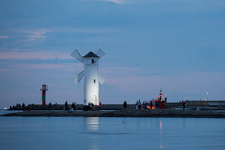 Tourists are walking near the Stawa Mlyny; a distinctive, historic navigational aid shaped like a white windmill, located at the end of the Western Breakwater in Swinoujscie. Swinoujscie is a port city located at the northwestern tip of Poland. It is a popular health resort that attracts tourists with its unique atmosphere and the widest natural beach in Poland.