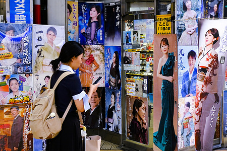 A woman uses her iPhone in front of a music shop specializing in enka singers in Shinagawa Ward, Tokyo. Promotional posters featuring enka performers are displayed outside the shop.
