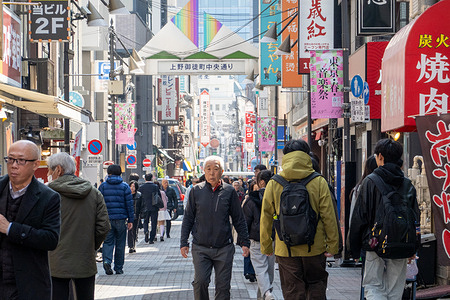 Pedestrians walk along a busy shopping street in the Ueno Okachimachi area of Tokyo, Japan. The district is known for its dense mix of restaurants, retail shops, and colorful signage attracting locals and visitors. The Ueno–Okachimachi area features busy shopping streets lined with discount stores, market stalls, and casual eateries catering to both locals and visitors. Centered around Ameya-Yokocho, the neighborhood has a lively, fast-paced atmosphere with a mix of street food, retail, and everyday city life.
