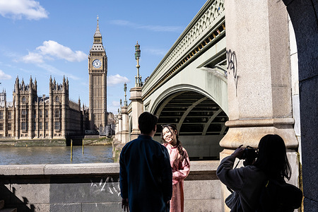 Foreign tourists seen posing for photos besides Westminster Bridge with a view of Big Ben and the Palace of Westminster in the background.