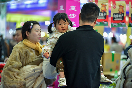 A father carries his young daughter on his back while a woman sits nearby in a colorful, crowded market.