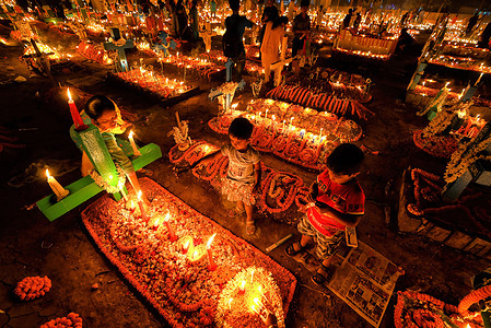 Kids offer prayers beside the grave of their deceased relative during All Souls' Day at a cemetery. Christian devotees gather at the graves of their departed loved ones at a cemetery with lit-up candles and flowers during All Souls' Day. This comes from an ancient belief that the souls of the dead will return, on this particular day, to have a meal with their family & friends. Candles are lit to guide the souls to their homes.