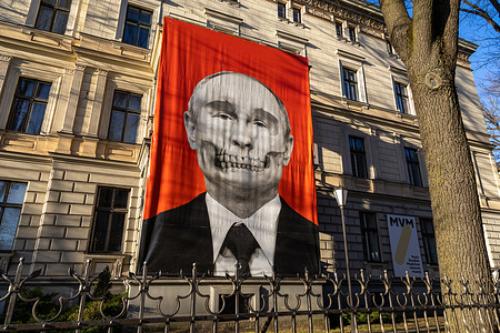 A giant protest banner depicting a skull-faced portrait of Vladimir Putin hangs on a building's facade near the Russian Embassy.