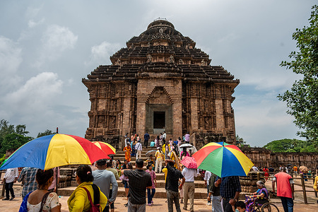 Tourists gather in the courtyard of the Konark Sun Temple and look toward the main structure, with several visitors carrying umbrellas under the strong sunlight in Konark, Puri. Designated as a UNESCO World Heritage Site, the 13th-century Konark Sun Temple was constructed around 1250 CE by King Narasimhadeva I of the Eastern Ganga dynasty. Built in the classic Kalinga architectural style, the complex forms a giant stone chariot for the Sun God, complete with 24 carved wheels and seven horses. While much of the original main sanctum has fallen into ruin, its surviving assembly hall (Jagamohana) remains a masterpiece of ancient Indian stonemasonry.