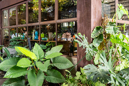 A close-up from the outside showing a customer sitting in the coffee shop and looking at his phone at Eastern Glass Manufacturer in Bangkok. Eastern Glass Manufacturer in Bangkok, with over 70 years of activity, is one of the last long-standing handmade glassware factories in the city, producing artisan pieces across generations. When international export orders dropped during COVID, the company adapted by opening a showroom and café within the original industrial structure to reach local customers and keep the factory active.