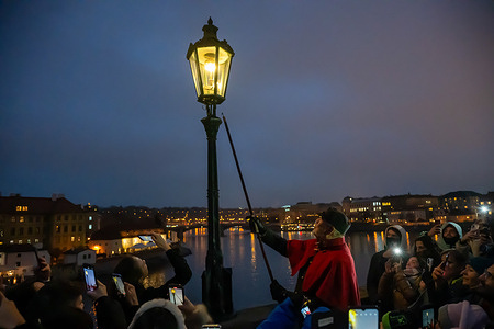 A lamplighter in a traditional red-and-black uniform lights a gas lamp on Prague’s iconic Charles Bridge as tourists look on. During the Advent season, the bridge’s lamps are lit manually instead of by the usual automated system. Prague’s gas street lighting tradition dates back to 1847.
