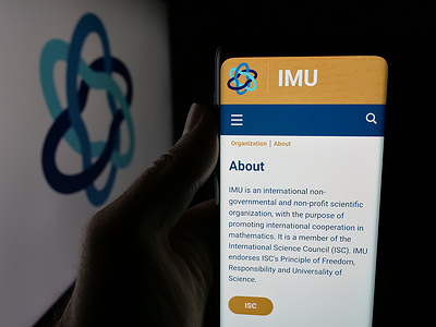 In this photo illustration, a person is holding a smartphone with the webpage of International Mathematical Union (IMU) on screen in front of logo.