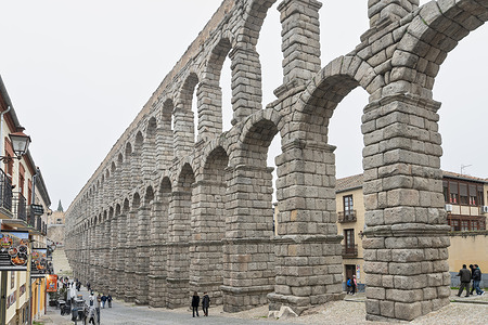 Daytime view of the Roman Aqueduct of Segovia, a UNESCO World Heritage Site in Spain. The well-preserved, double-tiered granite arches dominate the Plaza del Azoguejo, showcasing a massive feat of ancient engineering amidst a bustling modern city.