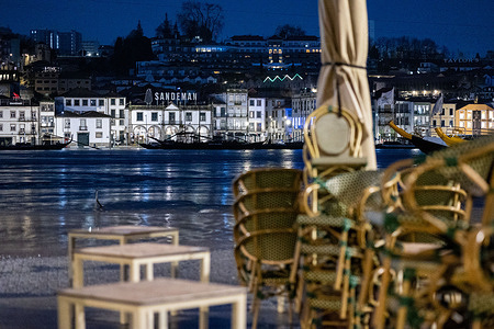Stacked café furniture seen beside the Douro as high tide raises water levels along the waterfront with reflections from illuminated buildings and boats shaping the night scene.