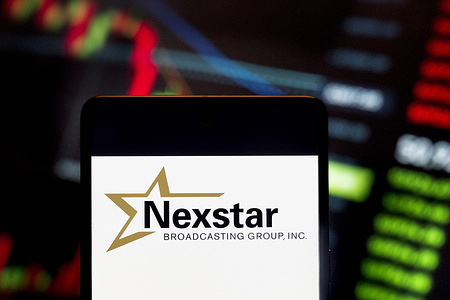 In this photo illustration, the American media company, Nexstar Media Group (NASDAQ: NXST), logo seen displayed on a smartphone with an economic stock exchange index graph in the background.