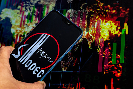 In this photo illustration, the Sinopec logo is seen displayed on a phone screen with a world map and stock market graph displayed on a computer screen.