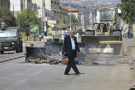 A Palestinian passes in front of an Israeli military bulldozer removing containers that were burned by Palestinian protesters that closed the street during a demonstration after Jewish settlers attacked them and killed a defenseless young man, Labib Muhammad, in the town of Howwarah.