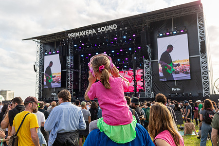 A young festivalgoer enjoys the atmosphere at Primavera Sound Porto, seated on someone's shoulders and wearing pink headphones to protect against loud music. The event, known for its diverse lineup and vibrant crowd, brings music lovers together for unforgettable experience.