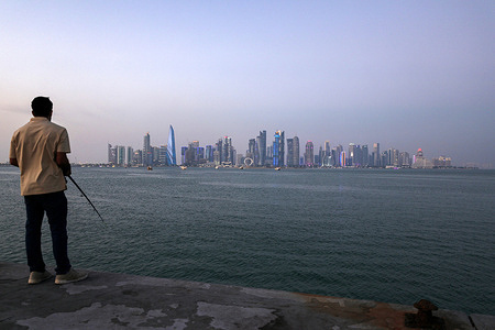 A man casts his fishing line into the sea, with towering skyscrapers on the Doha Corniche in the background.
