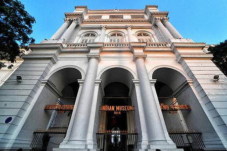 The entrance of the Indian Museum is shown with a metal gate and an orange sign reading 'INDIAN MUSEUM 1814' mounted above, framed by white neoclassical columns and an arched passageway.