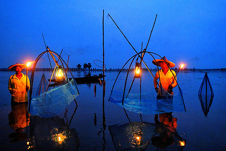 Fishermen seen hunting fish during the golden light hour of sunset using Hurricane Lamp Bortir Bill, a vast wetland surrounded by farmland in the North 24 Parganas district of West Bengal, about 50 kilometers from Kolkata, supports fishing as a primary source of livelihood for local residents. Despite the widespread use of modern fishing methods, many families continue to rely on traditional, low-cost, and environmentally friendly techniques, including bamboo fish traps, large fishing nets, and a distinctive leg-rowing method.