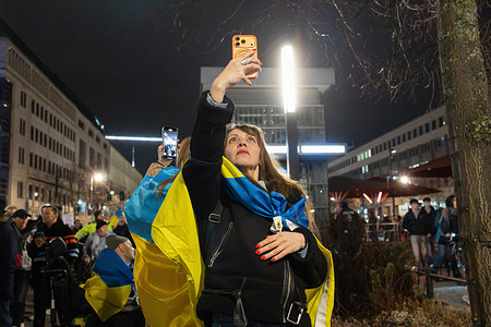A woman takes a selfie with an iPhone while holding a Ukrainian flag during the demonstration. A large demonstration were held in Berlin to mark the fourth anniversary of Russia’s full-scale war in Ukraine. Several thousand people marched from Lustgarten to the Brandenburg Gate in solidarity with Ukraine and to call for continued support, with police estimating about 5,000 participants. The march passed the Russian Embassy under heavy police presence, with participants carrying Ukrainian flags and anti-war signs. Speeches at the Brandenburg Gate included Ukraine’s ambassador to Germany, Oleksii Makeiev, and Berlin Mayor Kai Wegner, and the landmark was later lit in the colors of the Ukrainian flag.