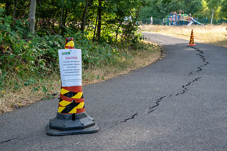 A traffic cone displaying a hazard warning sign seen placed at the start of a cracked path warning walkers and other road users of a potential hazard. After weeks of excessively hot and dry weather, some roads have begun to deteriorate with sections of the walkways cracking and breaking up. The city and Ewell Borough Council placed warning signs, cones and barrier tape around affected areas to alert walkers and other road users of the damage and potential hazards.