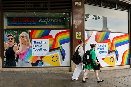 People walk past rainbow - colored window decorations at the Tesco Express celebrating Pride.