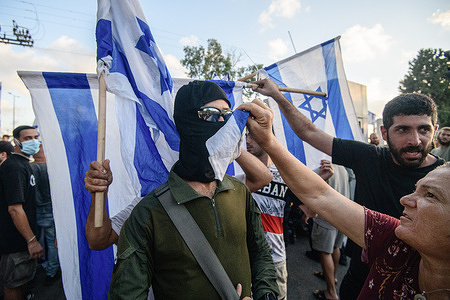 Supporting right-wing activists hide the face of an armed and masked reservist from the '100 force' with Israeli flags. Israeli right-wing activists demonstrated with masked IDF reservists from the ‘100 force’ and broke into ‘Beit Lid’ military police base following the detention of 9 military reservists from the unit today, who were suspected of sexual abuse of a detainee following the October 7 attack in Israel at the ‘Sde Teiman’ compound.