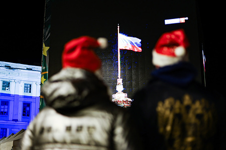 A couple wearing New Year's hats stand in front of a screen displaying the Russian flag during the New Year's celebrations.
