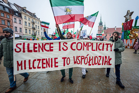 The march begins behind a banner that says, in English and Polish, "Silence is complicity" during the demonstration. Protesters of the Iranian diaspora gather in Warsaw, Poland to march from Castle Square to Staszic Palace. Following last month's anti-government protests in Iran, the demonstrators rally demanding an end to the violence of the Islamic Republic, and regime change to return the monarchy, which would install the Crown Prince of Iran, Reza Pahlavi, the son of the deposed Shah. The rally includes singing, chanting, dancing, and the waving of flags.
